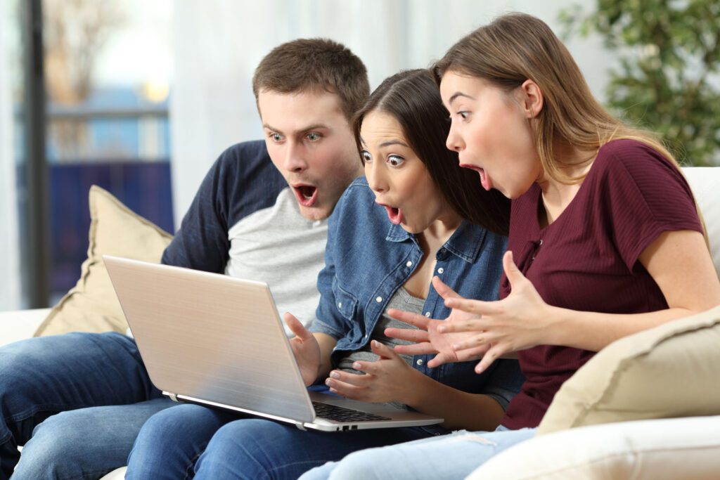 Three amazed friends watching media content on line in a computer sitting on a couch in the living room at home